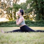 A woman performs a seated yoga pose outdoors in a serene meadow setting during daylight.